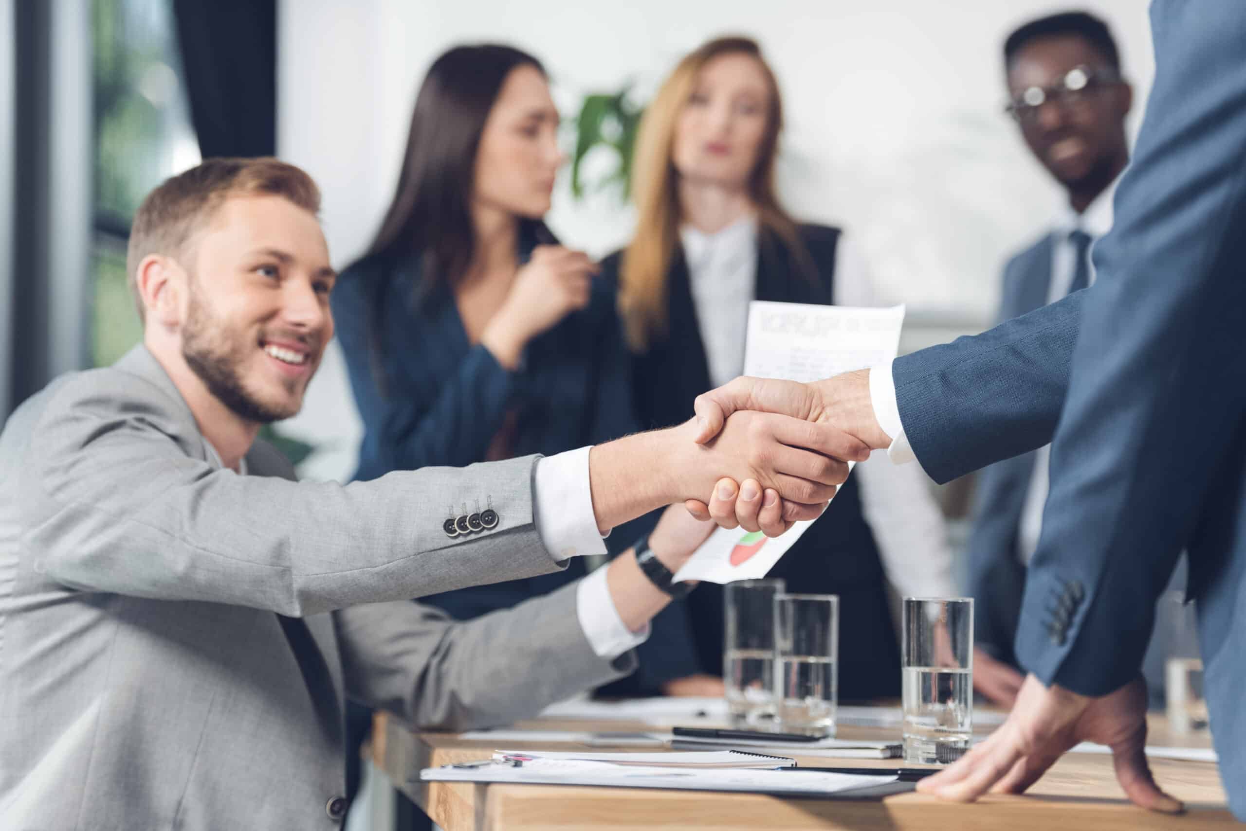 Two professionals handshake, symbolizing partnership in Seattle's IT services sector with colleagues in the background.